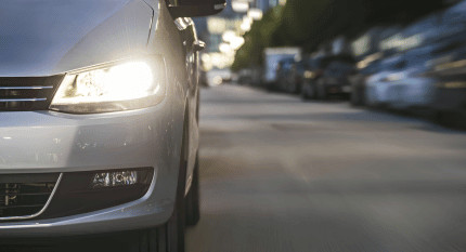 Faro delantero encendido de un coche gris circulando por una calle urbana al atardecer, con tráfico y coches aparcados al fondo.